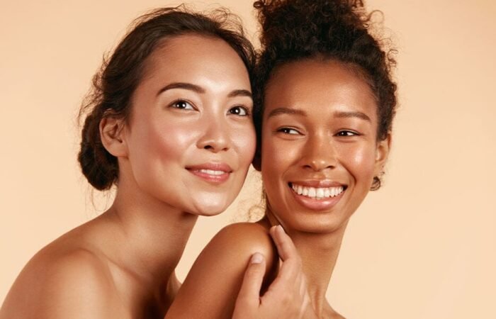An Asian woman and a Black woman with hair in buns smile into the distance with their heads pressed together in friendship.