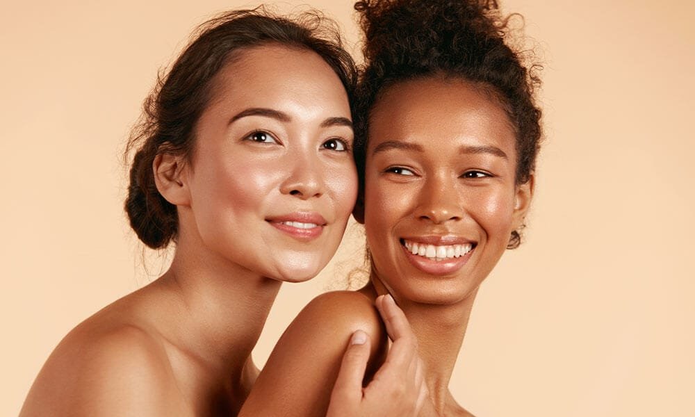 An Asian woman and a Black woman with hair in buns smile into the distance with their heads pressed together in friendship.